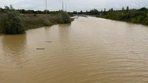 Tony Fisher/BBC The top of the Lower Shelton pumping station can be seen on the left, submerged in floodwater on the A421 near the bridge that carries Beancroft Road