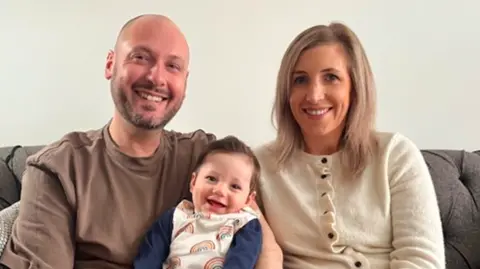 Group image of David and his wife Siobhan smiling as they hold baby Rupert betwen them. They are sat on a sofa with the smilng baby.