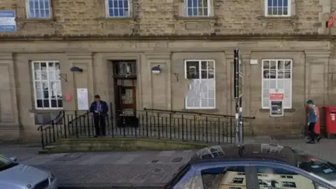 Frontage of Kendal Post Office, which is an old stone building with bay windows and a cash point. There are two red post boxes outside and people walk into the building.