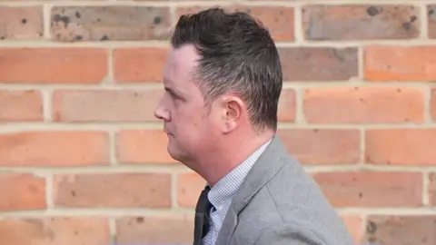 Simon Taylor outside Norwich Crown Court. He is walking and is looking away from the camera. He is wearing a grey suit and black tie. He is walking past a brick wall.