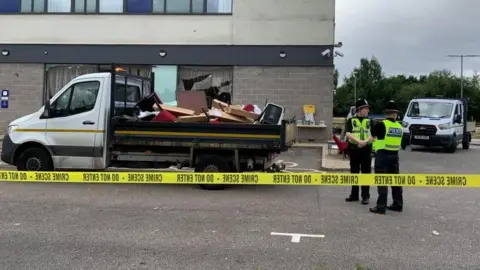 Two police officers stand in front of a building with broken windows, with crime scene tape sealing off the area.