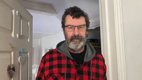 Man with dark curly hair and beard wearing a black and red checked jacket standing in a doorway