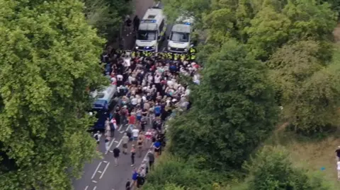 Essex Police An image taken by a drone, showing police officers using vans to block a large group of people from continuing down a road. The road is flanked by tall trees.