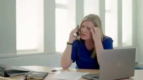 Cath Stanczyzyn at a blue top on a phone sitting in an office. There is an office in front of it, with a laptop to its left and windows behind it
