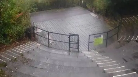 An amphitheatre in the rain. There is black metal fencing around the stage area with several layers of seating behind it, with steps down the centre.