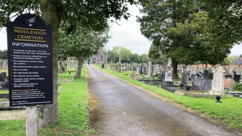 A view of Middlewich Cemetery. There is a line of gravestones on one side of a path, and several trees.