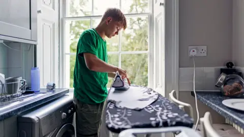 Getty Images Stock photo shows a person ironing a T shirt in front of a window in their kitchen, next to a plug socket and with the washing machine behind them.