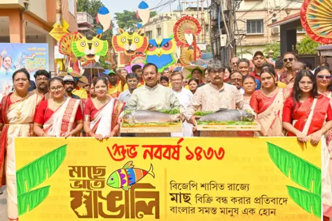 BBC A group of men and women wearing traditional attire campaign in Kolkata city pose for a photo. Two men at the centre are holding platters with big fish on them, while others around them hold colourful banners and placards