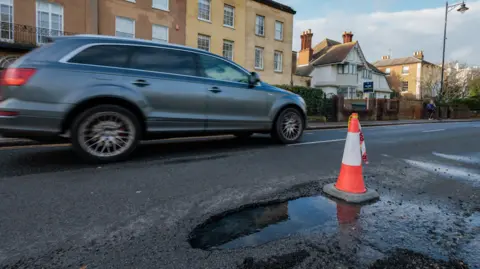 A car drives past a large pothole in the surface of a road that has fliled with water. Next to the pothole is a traffic cone. 