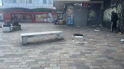 A stone bench in the foreground is surrounded by litter in front of a parade of shops including a vape shop and a fast food outlet, there's graffiti on shutters of a closed shop on the right hand side