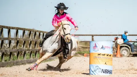 A girl wearing a cowboy hat, pink shirt and black trousers rides a grey horse at speed around and blue and yellow barrel, in a sandy arena surrounded by a fence