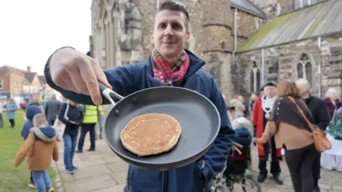 A man smiling holding a frying pan with a small pancake.