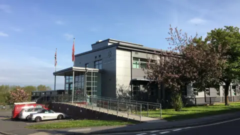 Several cars parked in front of a large grey building, which is the Shropshire Fire and Rescue Service headquarters, pictured from the side. Two flag poles with flags at the top are either side of the front entrance, with a ramp leading up to it. Two trees are visible on the right.