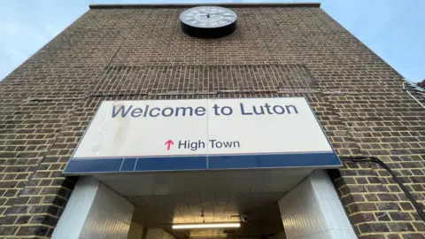 Ben Schofield/BBC Looking up an imposing brick wall, with a "welcome to Luton" sign hanging over a doorway, which is only partially visible. Higher above the sign is a clock, with a white face and Roman numerals.