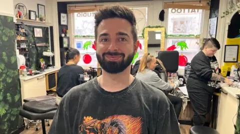 A smiling man with short spiky hair and a neatly-trimmed beard stands in a black t-shirt with an orange and red illustration on it. He stands at the front of a tattoo parlour with a tattoo artists and two clients behind him.