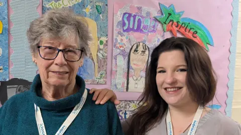 Tara Dolby/BBC Marvis Nicholson and Izzy Perks both smile at the camera as they stand side-by-side. Nicholson is wearing a green polo neck jumper, with glasses and has grey hair. Perks has long brown hair and is a wearing a grey blouse. They both have white lanyards around their necks. Behind them is a board of children's artwork. 