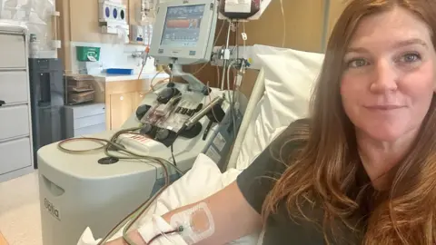 A woman with auburn hair sitting in a hospital bed. She has a needle in her right arm, which is pumping blood with the help of a large grey machine. Photo is a selfie.