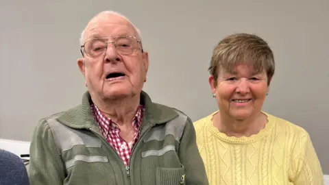 Jack Waterfield pictured with his daughter. Marilyn has short brunette hair and blue earrings, and is wearing a yellow knitted jumper. They are in front of a grey wall.