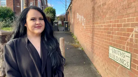 A woman with long black hair and a black blazer stands in front of an alleyway. There is a brick wall along the right one side of the alleyway and a row of houses along the other. There is a stone post in the middle of the alley and two wheelie bins further along. A street sign reads Smiths Buildings. 