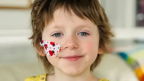 BBC Annabel, a young girl with blue eyes, short dark hair, and a tube coming from her nose concealed by a plaster with a ladybird pattern on it.
