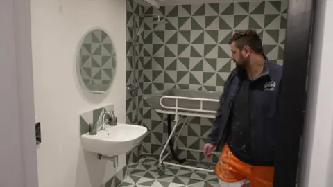 Lewis Franks stands on the right of the image looking towards a sink unit and behind them is a tiled walk in shower room