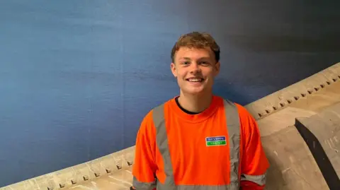 A young man with curly short blonde hair wearing a hi-vis jumper with Severn Trent written on it and standing in front of a picture of a dam.