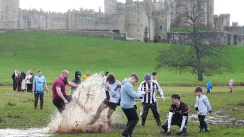 PA Media A group of players are surrounding the football. Several are turning away from a large spray of mud as one player kicks the ball. The castle can be seen in the background.