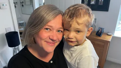 Kirsty is holding Harrison they are both smiling. She is wearing a black cardigan and he is wearing a white t-shirt. 