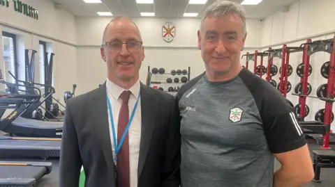 Two men stand side by side in a gym. On the left, a man is in a grey suit, with a white shirt and red tie. He wears glasses and has grey hair. On the right a man wears a grey T-shirt. He has grey hair. THere are treadmills and weights behind them.
