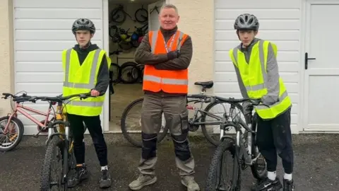 Travis, Danny and Thomas standing outside. They are wearing high vis-jackets and standing next to their bikes. Both Thomas and Travis are wearing a helmet.