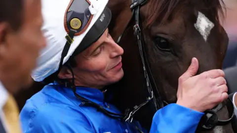 PA Media William Buick wears a white hat and blue silks and has his face next to a chestnut coloured horse with a white streak on it head. Buick's hand is on the animal's nose.
