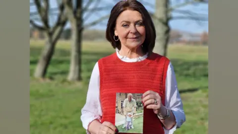 Carol Malia holding a photograph of her Dad Michael. She is wearing a red tank top and a white long-sleeved shirt and is standing in a sunny field with trees behind her.