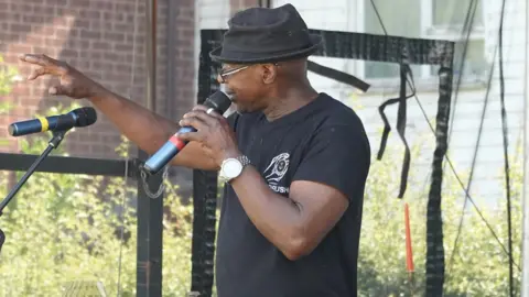 Ipswich Windrush Society A man in a trilby hat stands on stage with a microphone in his hand. One are is gesturing towards the crowd as he explains a point