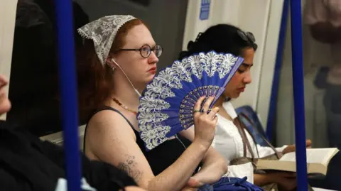 A woman with a bandana holding a blue fan sitting on the London underground