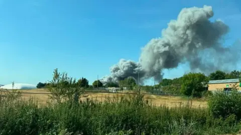 Naomi Pickett In the foreground is an arable field. In the distance, there is a large cloud of thick grey smoke rising into the blue, cloudless sky.