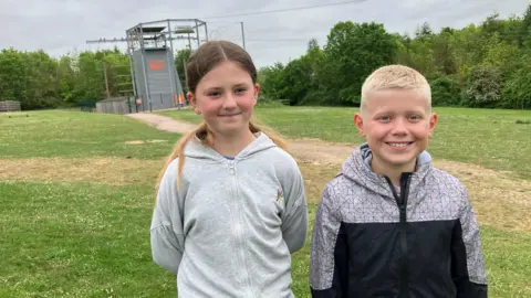 James Grant/BBC A girl in a grey hoodie and a boy in a grey and black top smile at the camera. A climbing wall is in the background.