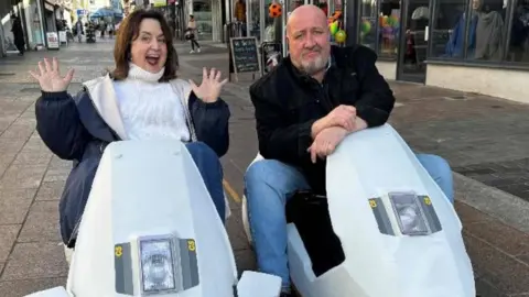 Ruth Jones and Steve Speirs on white scooters, Ruth has her hands in the air while Steve's hands are crossed and leaning on the scooter. They are parked in the middle of a street with shops on either side. 