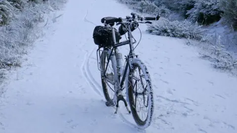 A snow covered bike without a rider on its stand on a snowy path