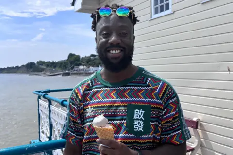 A man on Penarth Seafront holding an ice cream. He has sunglasses on his head and is wearing a colourful patterned t-shirt.