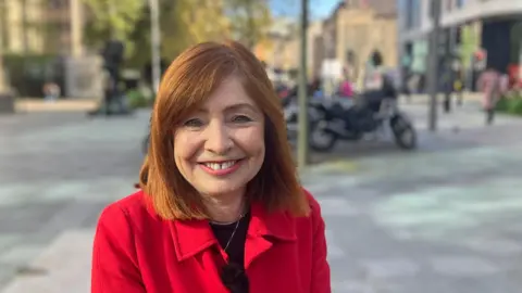 Sandra Busby, wearing a red coat, smiling at the camera in a street surrounded by office buildings