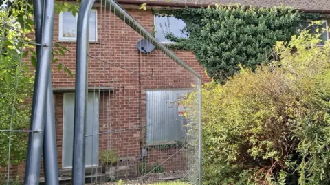 A close-up of a boarded up red-brick semi on The Brow, Huntingdon. Its ground floor door and window has metal shutters on it. Ivy is growing over the first floor window on the right. There is a metal fence in front of the property.