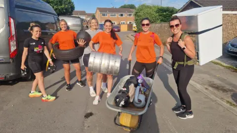 Commando Boot Camp Commando Boot Camp members pictured in orange t-shirts, smiling for the camera carrying fridges, tyres and weights in their hands 