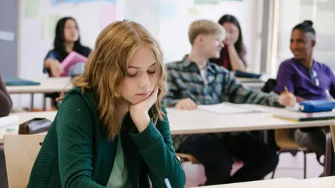 Getty Images Young students in classroom (stock photo). In the foreground is a young girl with shoulder-length blonde hair wearing a green jacket and top. He looks down and rests his face on his chin. Four other students are sitting in the background, two of whom are chatting.