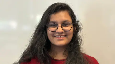 Une adolescente sourit à la caméra. Elle a de longs cheveux noirs et des lunettes et porte un haut rouge.