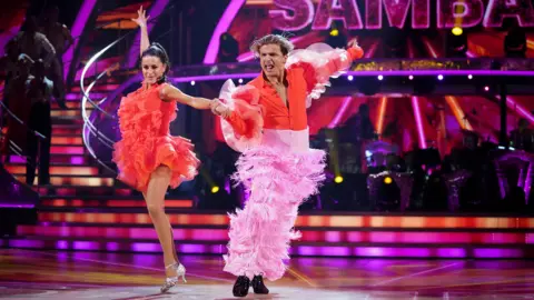 Amber and Nikita on the dance floor. Amber's one hand is straight in the air behind her mid dance move, while she holds Nikita's hand with the other. She is wearing a red frilled salsa dress and silver heels, while Nikita wears light pink tasselled trousers with a red shirt. His mouth is open and he also has an arm behind him, with the live band between them both. 