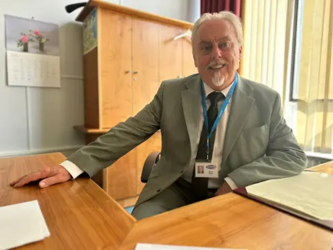 Robbie Roddis sits at a large wooden desk with a large wooden cabinet behind him. He has a grey/white goatee beard and grey hair and wears a grey suit with a black tie.
