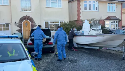 Police officers search the boot of a car and the open deck of a boat that are parked on a drive outside a house. An officer sits inside a police car in the foreground of the photo, the car being searched is in the middle of the frame, the boat is to the right. A neighbouring house can be seen on the other side of the boat