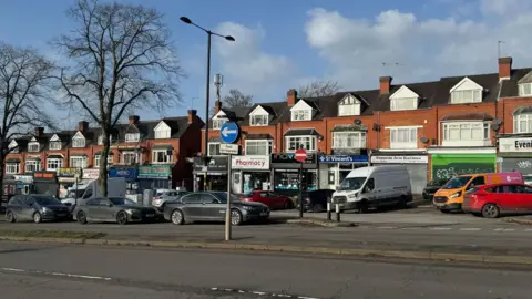 A row of shops and houses sit behind cars and vans.