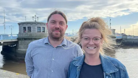 Andrew Turner/BBC Aaron Bennett, wearing a blue striped shirt. His hair is tied back, he is bearded and has a pierced lower lip. Georgia stands on the right. She has windblown blonde hair and wears glasses, a blue t-shirt and a blue denim jacket. They stand beside the River Yare in Great Yarmouth, with Haven Bridge in the background.