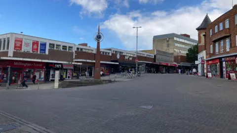 Pete Cooper/BBC A empty town centre with lots of high street shops and a thin red brick clock tower in the middle of the square. 